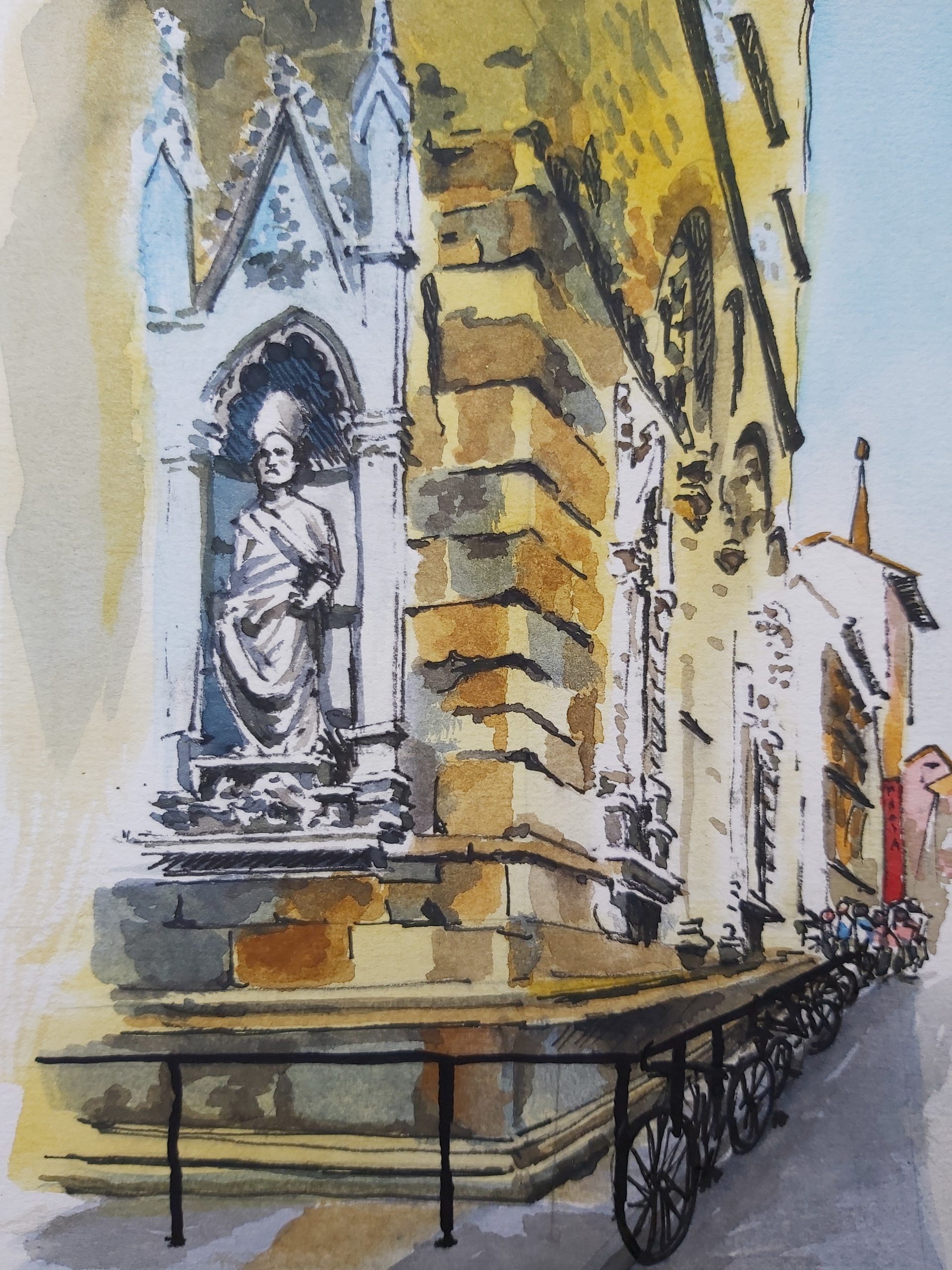 Watercolour image of a street corner in Florence featuring a religious statue set within a wall of a building.  A row of bicycles are laying against the railings outside. 